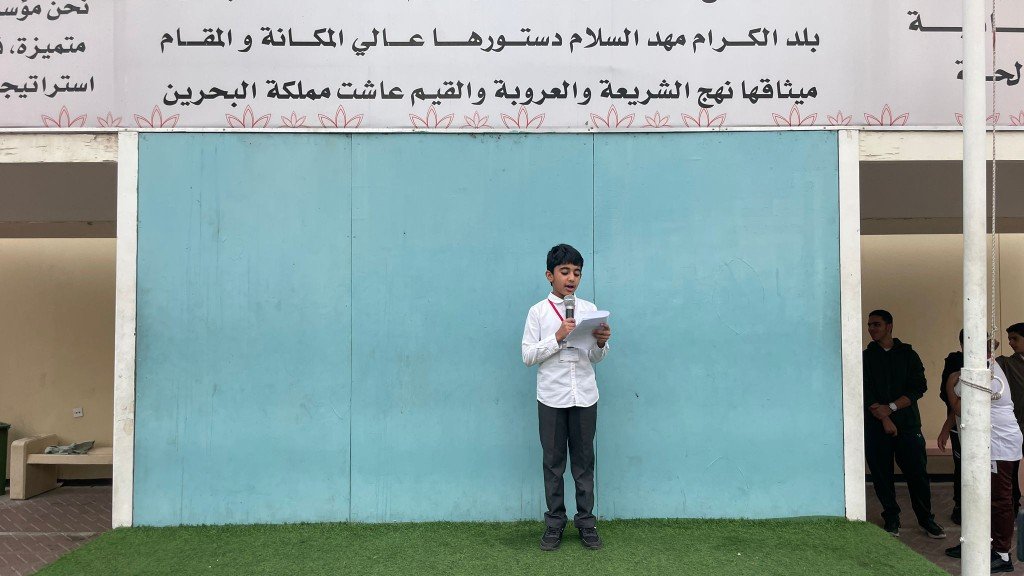 طالب بالزي المدرسي يتحدث بالمايكروفون على المنصة أمام خلفية المدرسة وشعار المملكة أثناء تفعيل الطابور الصباحي